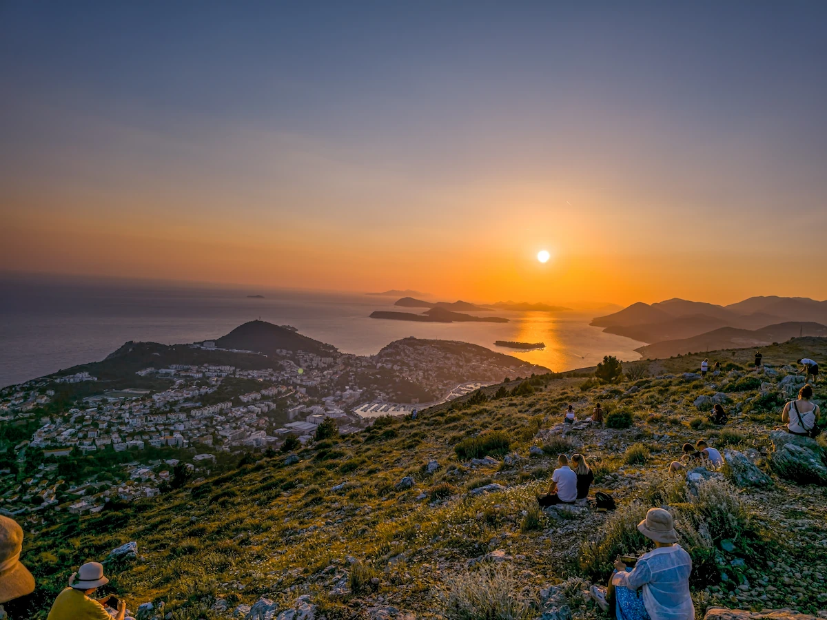 A wide golden hour panoramic view from the summit of Mount Srđ just before sunset, looking out over the calm Adriatic Sea and the glowing silhouettes of the Elaphiti Islands