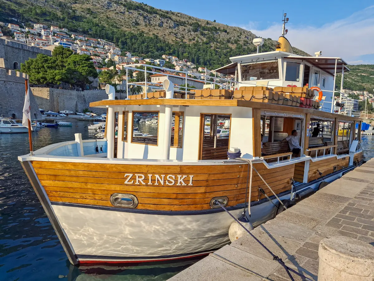 The wooden ferry boat Zrinski docked in the historic Dubrovnik Old Town harbor, preparing to depart for Lokrum Island